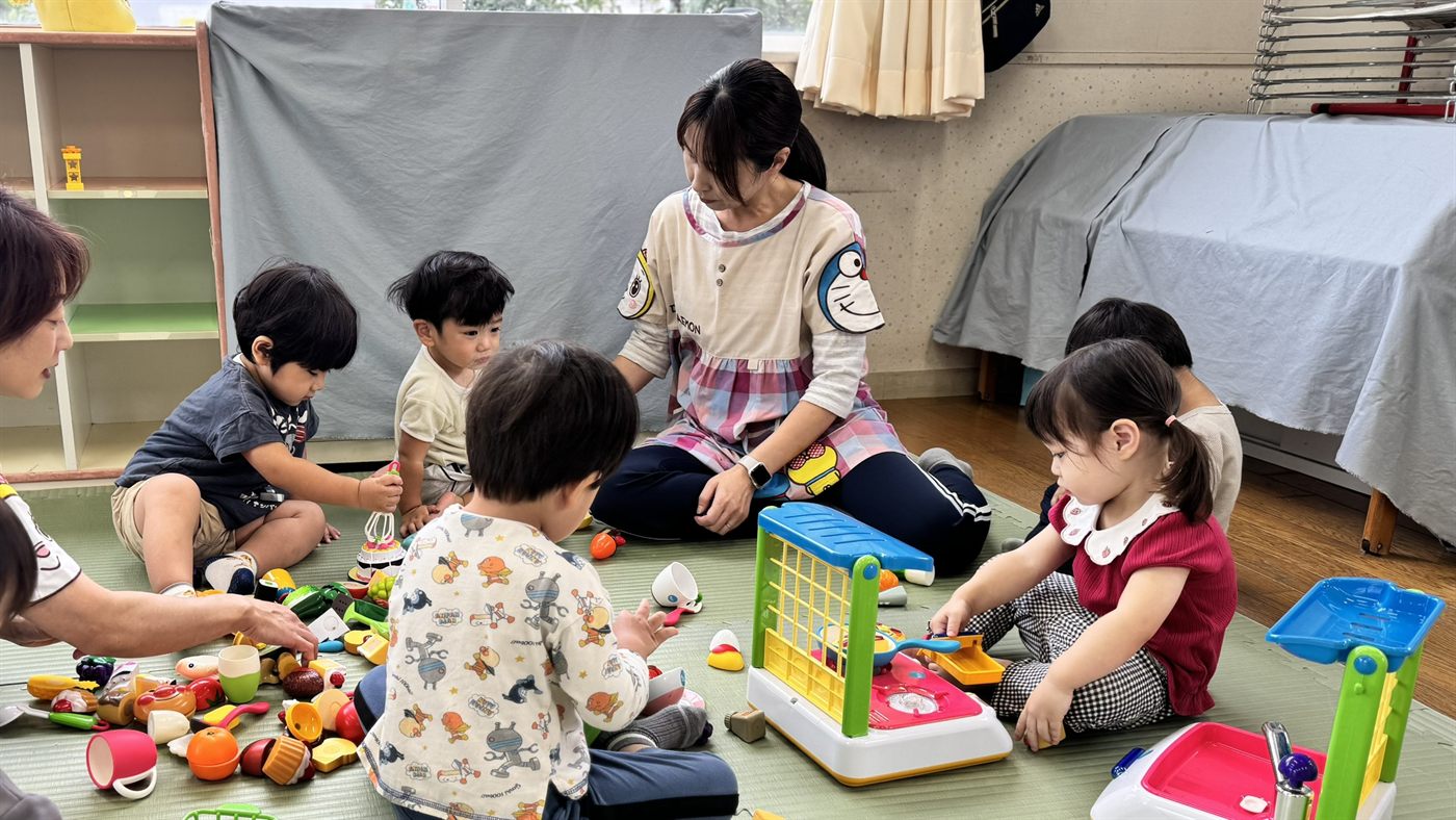 Children playing with toy kitchen sets alongside a teacher